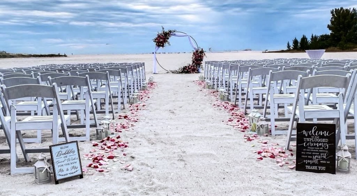 Beach ceremony setup with white chairs, rose petal aisle, and circular floral arch.