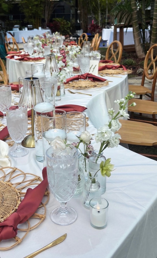 A long banquet table set with burgundy napkins, gold flatware, and white floral centerpieces under string lights.